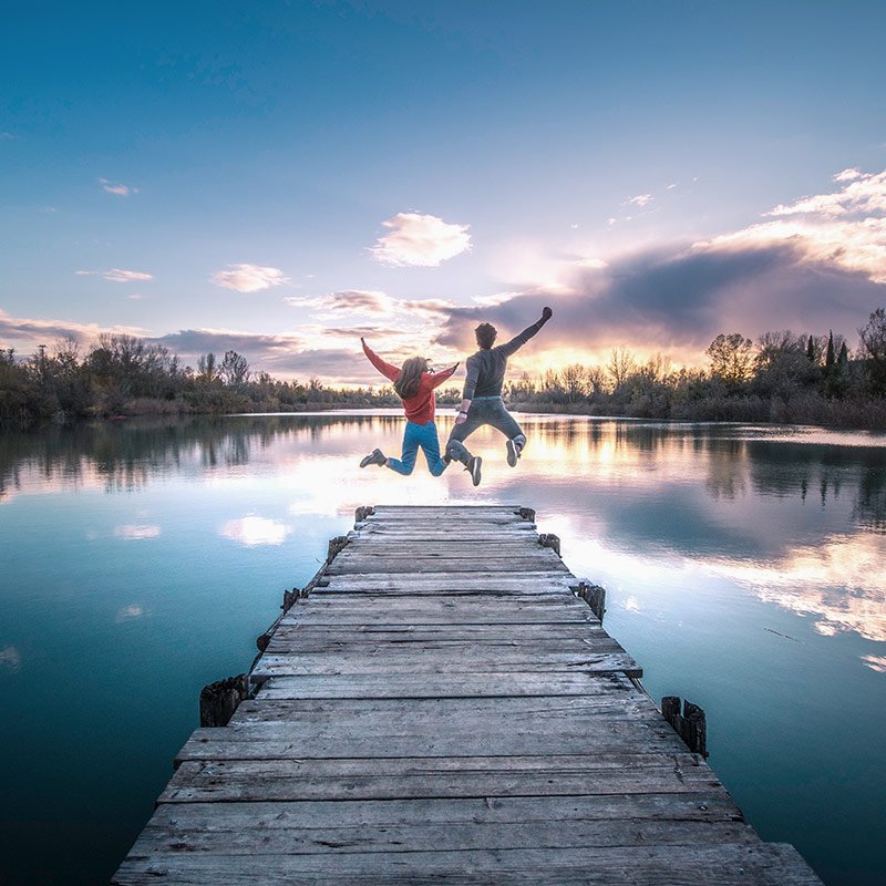 صديقان يقفزان فرحًا على رصيف خشبي عند بحيرة وقت الغروب